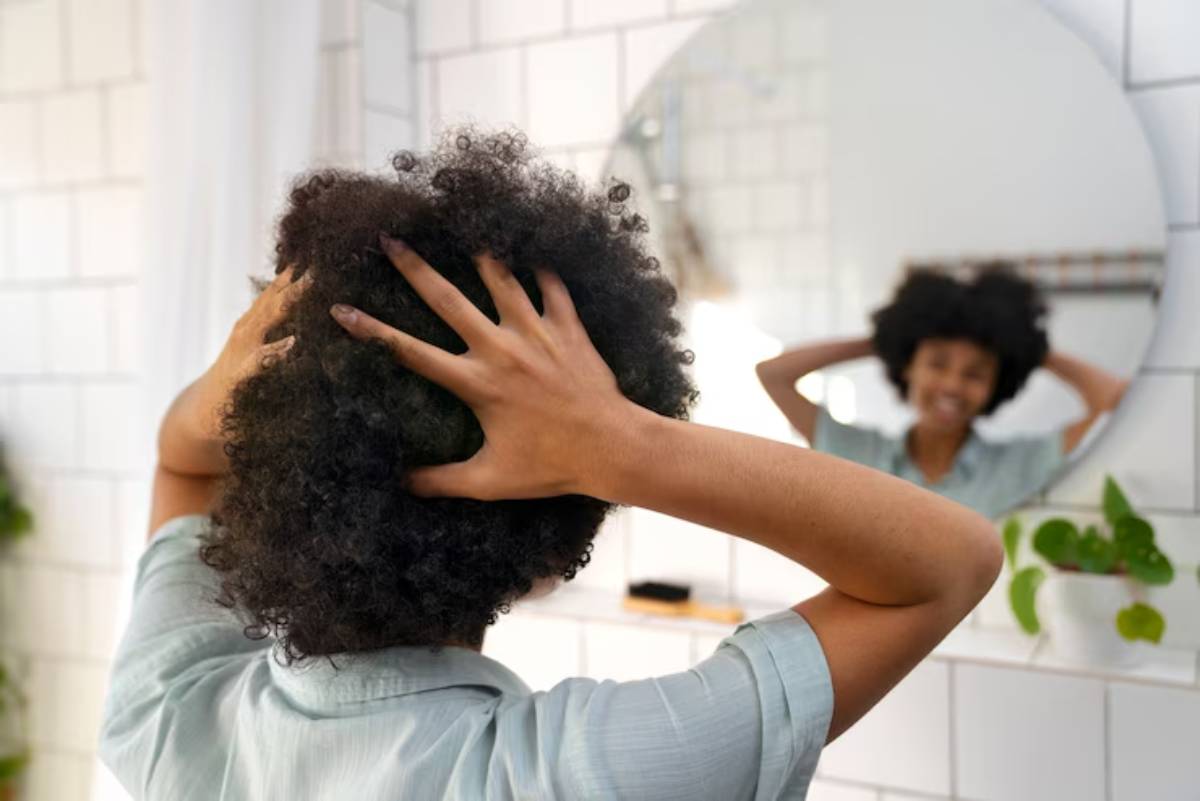 A person with curly hair is shown from behind, holding their head, in front of a circular bathroom mirror. Green plants are visible.