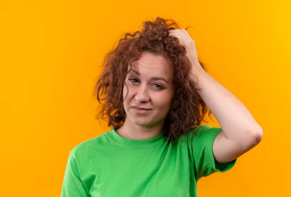  A person with curly hair in a green shirt gestures towards their head against a bright orange background.